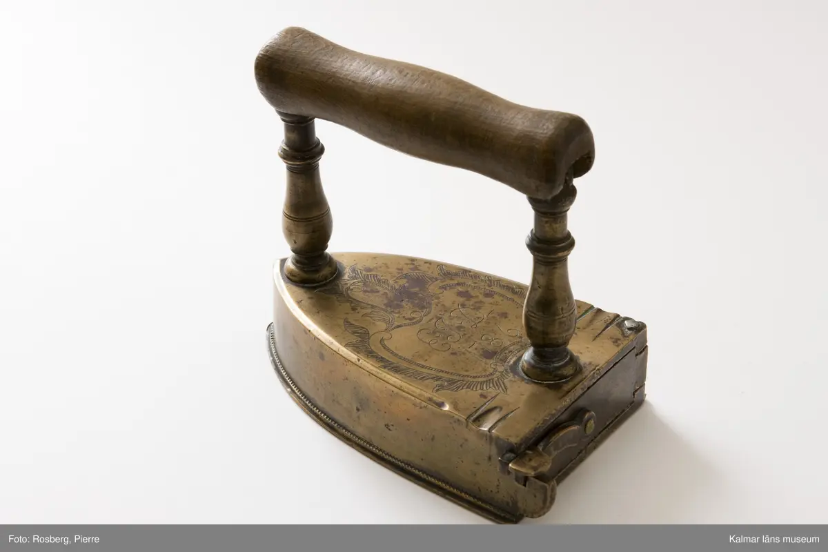 KLM 8511. Strykjärn. Tillverkat av malm eller mässing och trä. Handtag av trä. På järnets översida graverad ornering: EJD 1786 inom en rokokokartusch. Raka, profilerade stolpar till enkelt, runt handtag av trä.