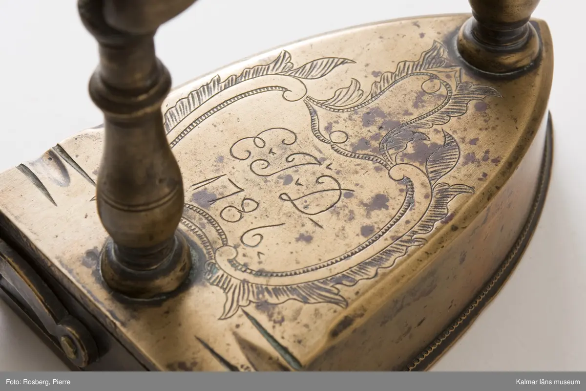 KLM 8511. Strykjärn. Tillverkat av malm eller mässing och trä. Handtag av trä. På järnets översida graverad ornering: EJD 1786 inom en rokokokartusch. Raka, profilerade stolpar till enkelt, runt handtag av trä.