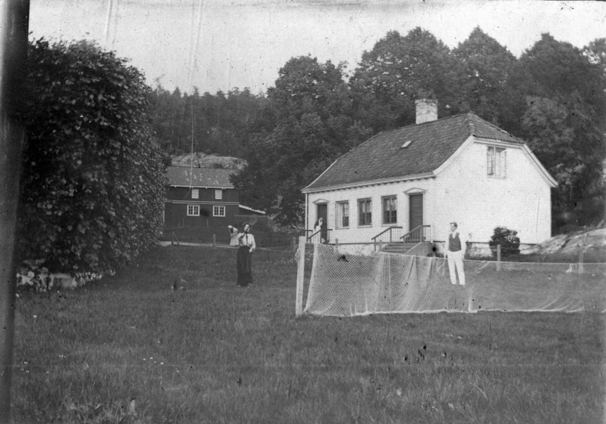Johanne Schweigaard og Sverre Heiberg spiller tennis. en kvinne står bak Johanne ved fjøset. En kvinne med forkle står ved ei dør til et av husene på Rørvik gård. ca. 1900