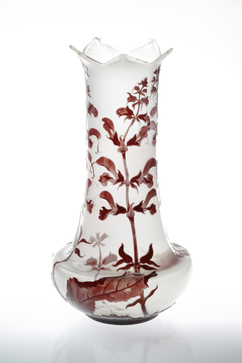 Høyreist vase i opakt melkehvitt glass. Nedre del av korpus er skålformet, hvorfra det reiser seg en høy sylinderformet hals. Vasens munning har en bladlignende utforming. Korpus er dekorert med overfangsteknikk, utformet som blomster og bladverk i lilla og burgunderrød. Dekoren er delvis bemalt med emaljemaling.