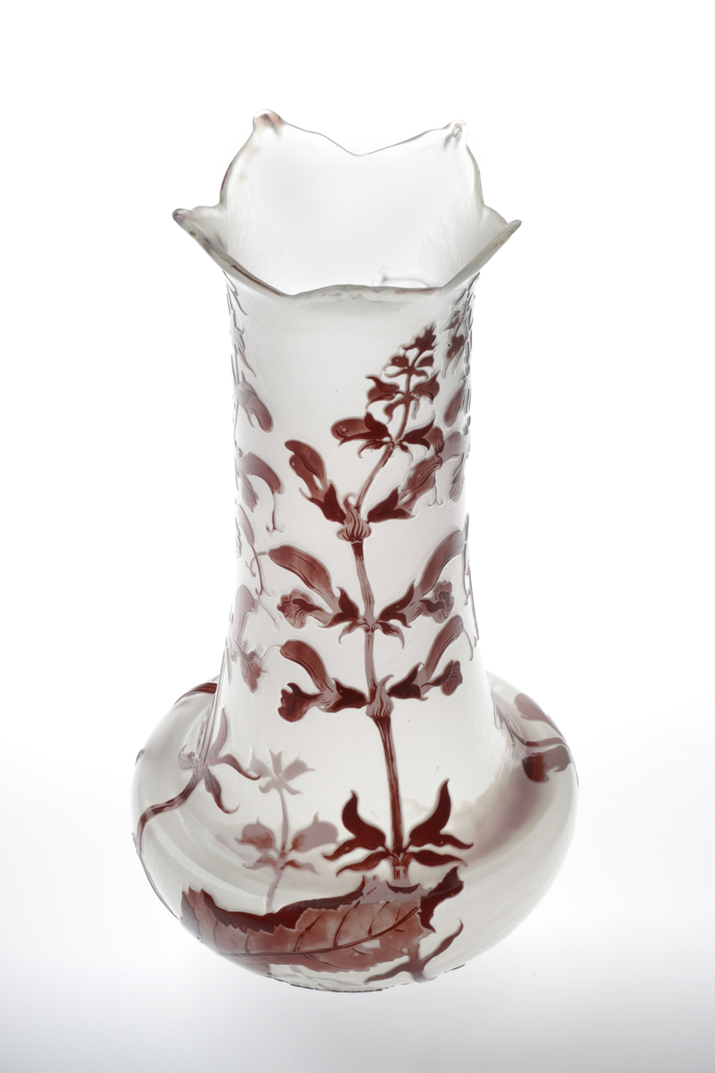 Høyreist vase i opakt melkehvitt glass. Nedre del av korpus er skålformet, hvorfra det reiser seg en høy sylinderformet hals. Vasens munning har en bladlignende utforming. Korpus er dekorert med overfangsteknikk, utformet som blomster og bladverk i lilla og burgunderrød. Dekoren er delvis bemalt med emaljemaling.
