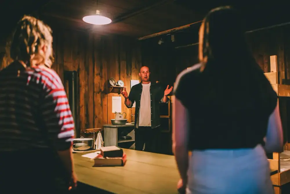 Fotografi i brakkerommet. En mann snakker og gestikulerer med hendene, han har ansiktet vendt mot kamera og mot to besøkende. Det er to kvinner som vi kun ser ryggen på.