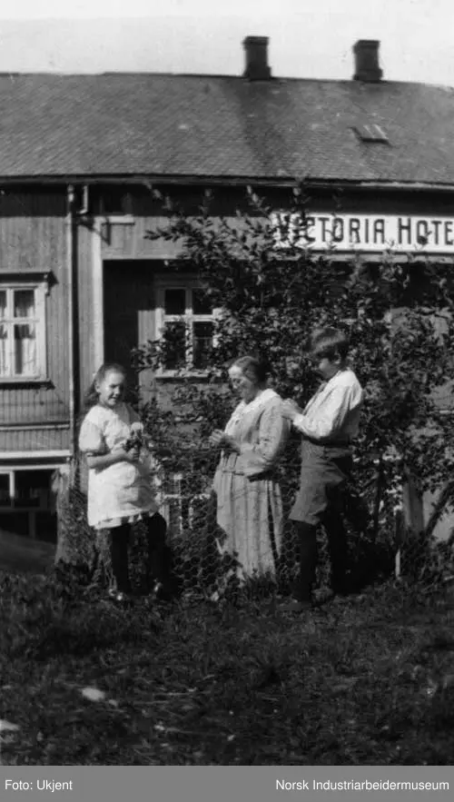 Tre mennesker stående i hagen utenfor Tvetens kafe i Telegata. Victoria hotell på andre siden av gaten.