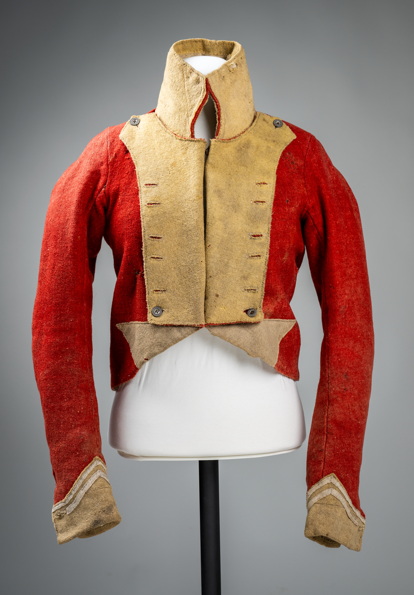 Kort trøye med høy ståkrage, sydd i grovt rødt lett valket ullstoff i empiresnitt. Trøya er helforet med grovt ullstoff. Kragen har mellomfor som er sydd fast til innerkragen. Ytterstoffet på kragen er brettet over på innsiden i øvre kant. Tittekant med rødt stoff i kortsidene på kragen. Trøya har søm MB og bakovertrekte skulder- og sidesømmer. Sidesømmene buer fra ermrigningen og mot MB. Sidesømmene slutter som legg med innlegg av gult stoff. Samme stoff brukes også som ytterkrage og slag på framstykket. Slagene er fastsydd og går opp som trekantforer mot kragen. Kneppes fast i bolen. 7 knapphull, men det mangler mange knapper. Plagget lukkes med 8 par metallhekter MF.  Nedenfor slagene er det påsydd trekanter av gråbeige ullstoff. Fra slagene foran skrår trøya bakover. Tosømsermer med dekor frampå ermene. Påsydd gult stoff med 2 striper av lys gråhvite bånd som går opp i en spiss mot innersømmen på ermet. Splitt ved yttersømmen. Denne lukkes med trekt rød knapp og knapphull. Mangler knappen på venstre erme. Stikninger langs slagene og kragen. Trøya er helt håndsydd og ytterkantene er sydd med sårkant på både for og stoff Tekst på 1 knapp, 3 knapper med stripe effekt inn mot midten.
Rød stoffhempe i nakken MB. Trøya har trolig tilhørt militæruniform fra tidlig 1800tall.