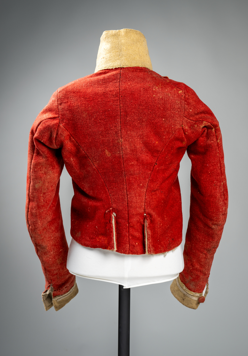 Kort trøye med høy ståkrage, sydd i grovt rødt lett valket ullstoff i empiresnitt. Trøya er helforet med grovt ullstoff. Kragen har mellomfor som er sydd fast til innerkragen. Ytterstoffet på kragen er brettet over på innsiden i øvre kant. Tittekant med rødt stoff i kortsidene på kragen. Trøya har søm MB og bakovertrekte skulder- og sidesømmer. Sidesømmene buer fra ermrigningen og mot MB. Sidesømmene slutter som legg med innlegg av gult stoff. Samme stoff brukes også som ytterkrage og slag på framstykket. Slagene er fastsydd og går opp som trekantforer mot kragen. Kneppes fast i bolen. 7 knapphull, men det mangler mange knapper. Plagget lukkes med 8 par metallhekter MF.  Nedenfor slagene er det påsydd trekanter av gråbeige ullstoff. Fra slagene foran skrår trøya bakover. Tosømsermer med dekor frampå ermene. Påsydd gult stoff med 2 striper av lys gråhvite bånd som går opp i en spiss mot innersømmen på ermet. Splitt ved yttersømmen. Denne lukkes med trekt rød knapp og knapphull. Mangler knappen på venstre erme. Stikninger langs slagene og kragen. Trøya er helt håndsydd og ytterkantene er sydd med sårkant på både for og stoff Tekst på 1 knapp, 3 knapper med stripe effekt inn mot midten.
Rød stoffhempe i nakken MB. Trøya har trolig tilhørt militæruniform fra tidlig 1800tall.