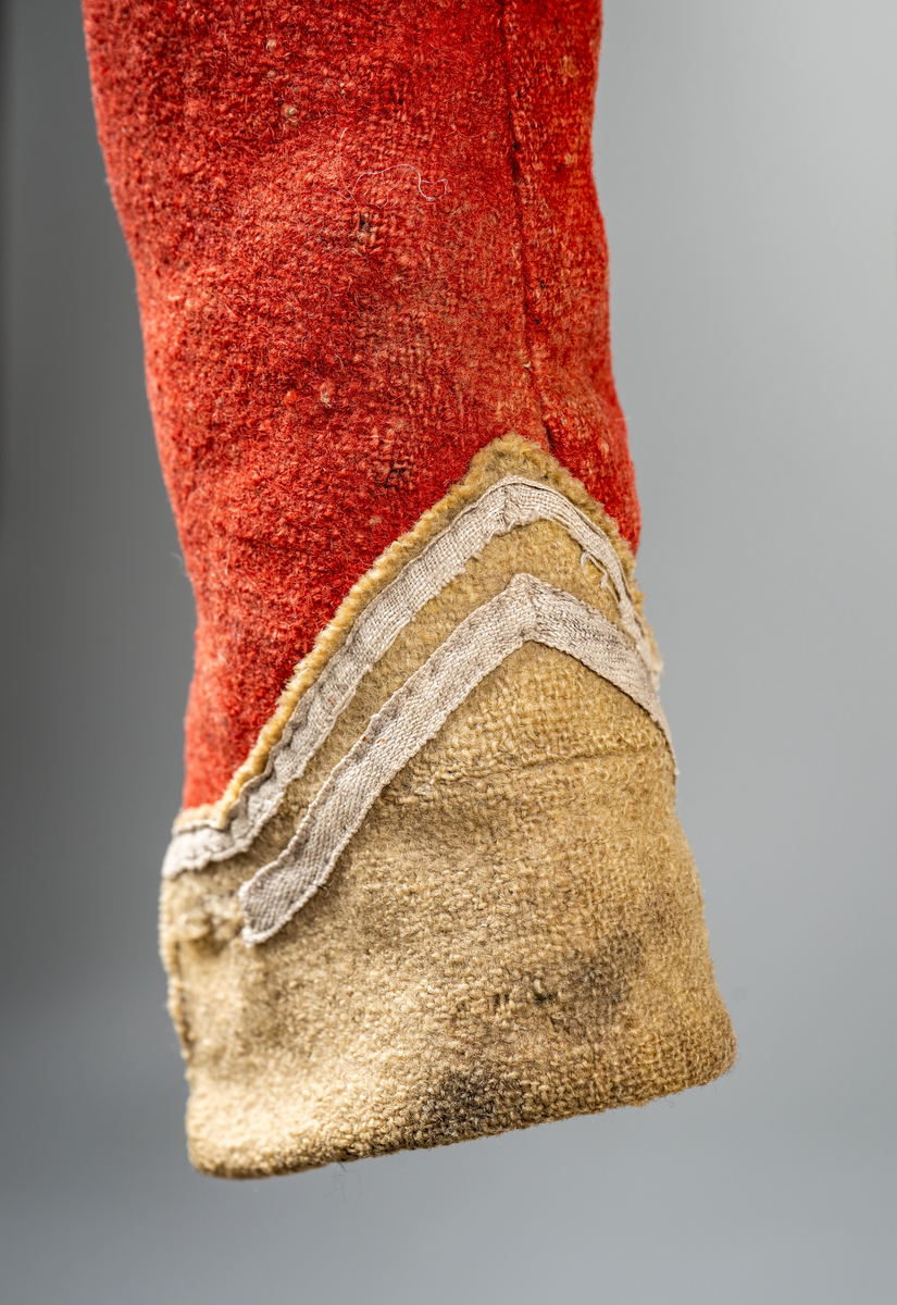 Kort trøye med høy ståkrage, sydd i grovt rødt lett valket ullstoff i empiresnitt. Trøya er helforet med grovt ullstoff. Kragen har mellomfor som er sydd fast til innerkragen. Ytterstoffet på kragen er brettet over på innsiden i øvre kant. Tittekant med rødt stoff i kortsidene på kragen. Trøya har søm MB og bakovertrekte skulder- og sidesømmer. Sidesømmene buer fra ermrigningen og mot MB. Sidesømmene slutter som legg med innlegg av gult stoff. Samme stoff brukes også som ytterkrage og slag på framstykket. Slagene er fastsydd og går opp som trekantforer mot kragen. Kneppes fast i bolen. 7 knapphull, men det mangler mange knapper. Plagget lukkes med 8 par metallhekter MF.  Nedenfor slagene er det påsydd trekanter av gråbeige ullstoff. Fra slagene foran skrår trøya bakover. Tosømsermer med dekor frampå ermene. Påsydd gult stoff med 2 striper av lys gråhvite bånd som går opp i en spiss mot innersømmen på ermet. Splitt ved yttersømmen. Denne lukkes med trekt rød knapp og knapphull. Mangler knappen på venstre erme. Stikninger langs slagene og kragen. Trøya er helt håndsydd og ytterkantene er sydd med sårkant på både for og stoff Tekst på 1 knapp, 3 knapper med stripe effekt inn mot midten.
Rød stoffhempe i nakken MB. Trøya har trolig tilhørt militæruniform fra tidlig 1800tall.