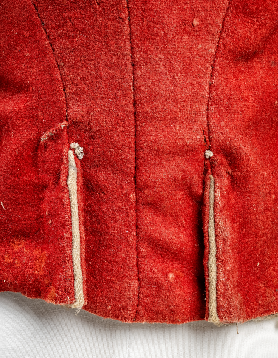 Kort trøye med høy ståkrage, sydd i grovt rødt lett valket ullstoff i empiresnitt. Trøya er helforet med grovt ullstoff. Kragen har mellomfor som er sydd fast til innerkragen. Ytterstoffet på kragen er brettet over på innsiden i øvre kant. Tittekant med rødt stoff i kortsidene på kragen. Trøya har søm MB og bakovertrekte skulder- og sidesømmer. Sidesømmene buer fra ermrigningen og mot MB. Sidesømmene slutter som legg med innlegg av gult stoff. Samme stoff brukes også som ytterkrage og slag på framstykket. Slagene er fastsydd og går opp som trekantforer mot kragen. Kneppes fast i bolen. 7 knapphull, men det mangler mange knapper. Plagget lukkes med 8 par metallhekter MF.  Nedenfor slagene er det påsydd trekanter av gråbeige ullstoff. Fra slagene foran skrår trøya bakover. Tosømsermer med dekor frampå ermene. Påsydd gult stoff med 2 striper av lys gråhvite bånd som går opp i en spiss mot innersømmen på ermet. Splitt ved yttersømmen. Denne lukkes med trekt rød knapp og knapphull. Mangler knappen på venstre erme. Stikninger langs slagene og kragen. Trøya er helt håndsydd og ytterkantene er sydd med sårkant på både for og stoff Tekst på 1 knapp, 3 knapper med stripe effekt inn mot midten.
Rød stoffhempe i nakken MB. Trøya har trolig tilhørt militæruniform fra tidlig 1800tall.