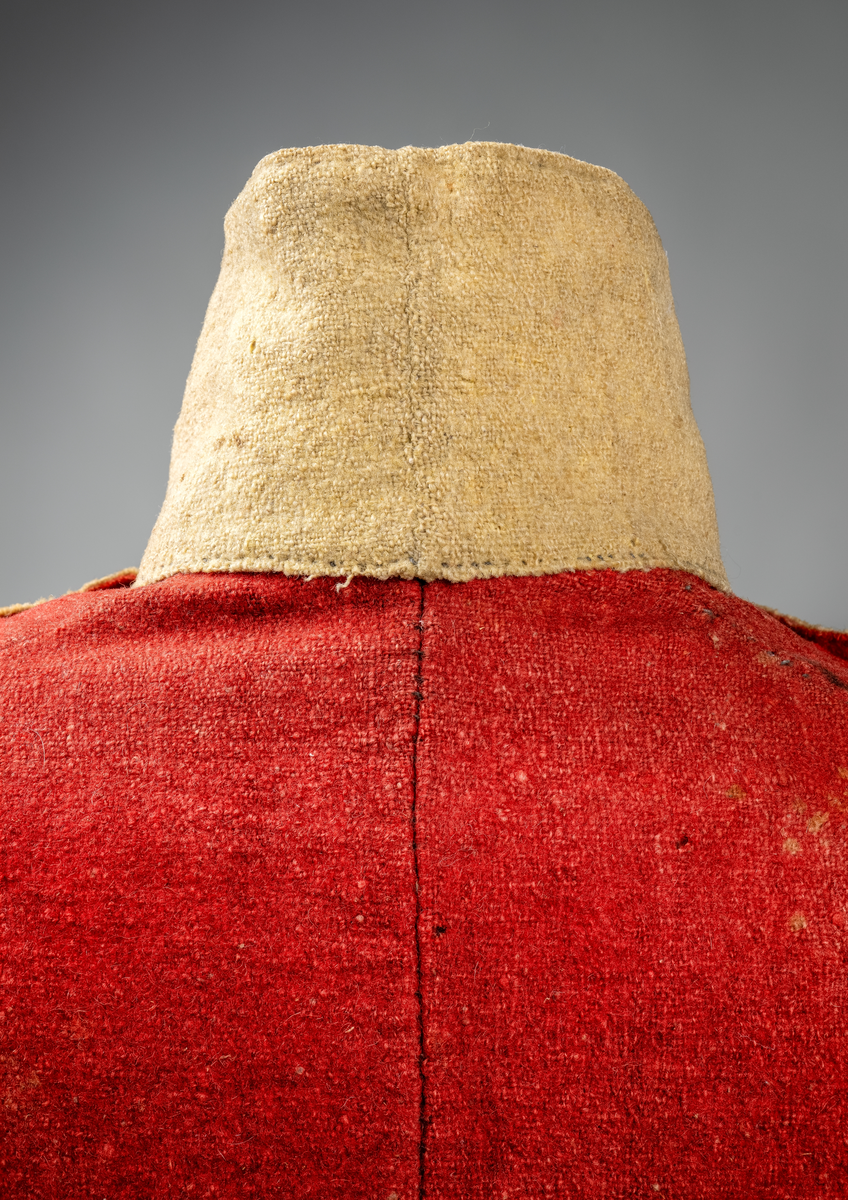 Kort trøye med høy ståkrage, sydd i grovt rødt lett valket ullstoff i empiresnitt. Trøya er helforet med grovt ullstoff. Kragen har mellomfor som er sydd fast til innerkragen. Ytterstoffet på kragen er brettet over på innsiden i øvre kant. Tittekant med rødt stoff i kortsidene på kragen. Trøya har søm MB og bakovertrekte skulder- og sidesømmer. Sidesømmene buer fra ermrigningen og mot MB. Sidesømmene slutter som legg med innlegg av gult stoff. Samme stoff brukes også som ytterkrage og slag på framstykket. Slagene er fastsydd og går opp som trekantforer mot kragen. Kneppes fast i bolen. 7 knapphull, men det mangler mange knapper. Plagget lukkes med 8 par metallhekter MF.  Nedenfor slagene er det påsydd trekanter av gråbeige ullstoff. Fra slagene foran skrår trøya bakover. Tosømsermer med dekor frampå ermene. Påsydd gult stoff med 2 striper av lys gråhvite bånd som går opp i en spiss mot innersømmen på ermet. Splitt ved yttersømmen. Denne lukkes med trekt rød knapp og knapphull. Mangler knappen på venstre erme. Stikninger langs slagene og kragen. Trøya er helt håndsydd og ytterkantene er sydd med sårkant på både for og stoff Tekst på 1 knapp, 3 knapper med stripe effekt inn mot midten.
Rød stoffhempe i nakken MB. Trøya har trolig tilhørt militæruniform fra tidlig 1800tall.