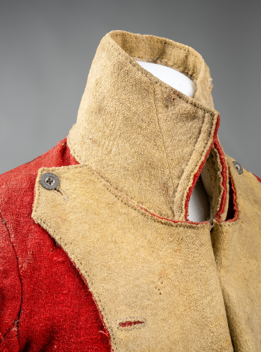 Kort trøye med høy ståkrage, sydd i grovt rødt lett valket ullstoff i empiresnitt. Trøya er helforet med grovt ullstoff. Kragen har mellomfor som er sydd fast til innerkragen. Ytterstoffet på kragen er brettet over på innsiden i øvre kant. Tittekant med rødt stoff i kortsidene på kragen. Trøya har søm MB og bakovertrekte skulder- og sidesømmer. Sidesømmene buer fra ermrigningen og mot MB. Sidesømmene slutter som legg med innlegg av gult stoff. Samme stoff brukes også som ytterkrage og slag på framstykket. Slagene er fastsydd og går opp som trekantforer mot kragen. Kneppes fast i bolen. 7 knapphull, men det mangler mange knapper. Plagget lukkes med 8 par metallhekter MF.  Nedenfor slagene er det påsydd trekanter av gråbeige ullstoff. Fra slagene foran skrår trøya bakover. Tosømsermer med dekor frampå ermene. Påsydd gult stoff med 2 striper av lys gråhvite bånd som går opp i en spiss mot innersømmen på ermet. Splitt ved yttersømmen. Denne lukkes med trekt rød knapp og knapphull. Mangler knappen på venstre erme. Stikninger langs slagene og kragen. Trøya er helt håndsydd og ytterkantene er sydd med sårkant på både for og stoff Tekst på 1 knapp, 3 knapper med stripe effekt inn mot midten.
Rød stoffhempe i nakken MB. Trøya har trolig tilhørt militæruniform fra tidlig 1800tall.