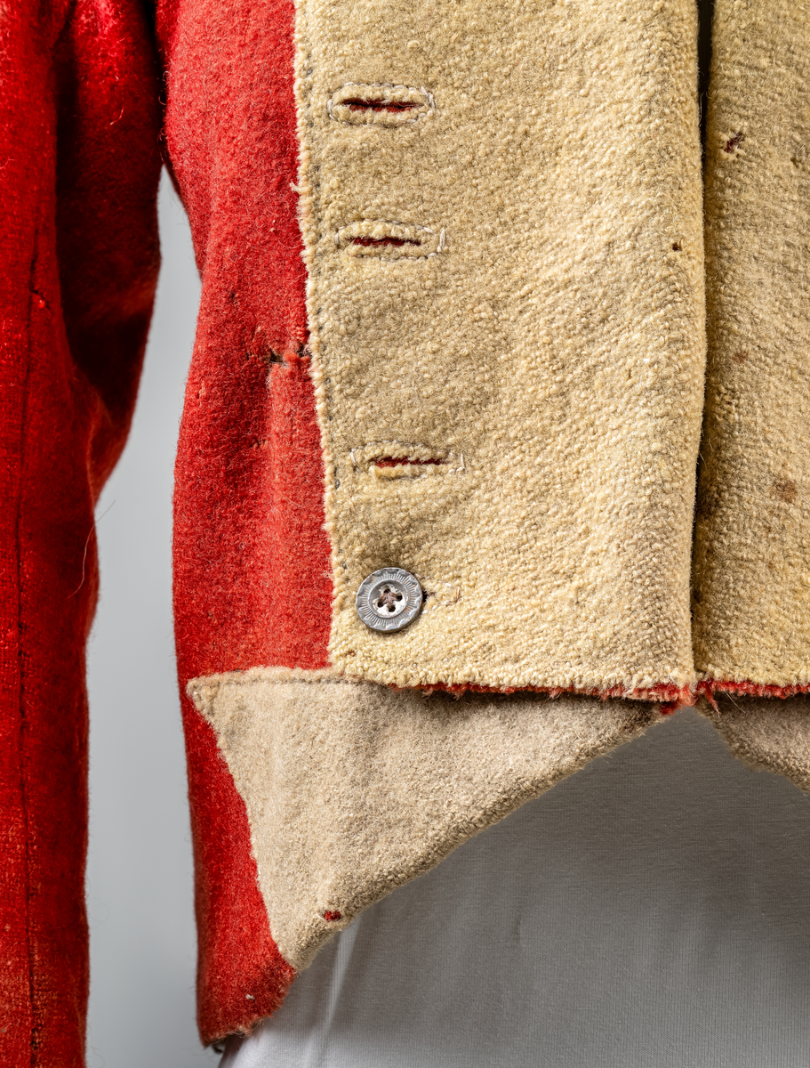 Kort trøye med høy ståkrage, sydd i grovt rødt lett valket ullstoff i empiresnitt. Trøya er helforet med grovt ullstoff. Kragen har mellomfor som er sydd fast til innerkragen. Ytterstoffet på kragen er brettet over på innsiden i øvre kant. Tittekant med rødt stoff i kortsidene på kragen. Trøya har søm MB og bakovertrekte skulder- og sidesømmer. Sidesømmene buer fra ermrigningen og mot MB. Sidesømmene slutter som legg med innlegg av gult stoff. Samme stoff brukes også som ytterkrage og slag på framstykket. Slagene er fastsydd og går opp som trekantforer mot kragen. Kneppes fast i bolen. 7 knapphull, men det mangler mange knapper. Plagget lukkes med 8 par metallhekter MF.  Nedenfor slagene er det påsydd trekanter av gråbeige ullstoff. Fra slagene foran skrår trøya bakover. Tosømsermer med dekor frampå ermene. Påsydd gult stoff med 2 striper av lys gråhvite bånd som går opp i en spiss mot innersømmen på ermet. Splitt ved yttersømmen. Denne lukkes med trekt rød knapp og knapphull. Mangler knappen på venstre erme. Stikninger langs slagene og kragen. Trøya er helt håndsydd og ytterkantene er sydd med sårkant på både for og stoff Tekst på 1 knapp, 3 knapper med stripe effekt inn mot midten.
Rød stoffhempe i nakken MB. Trøya har trolig tilhørt militæruniform fra tidlig 1800tall.