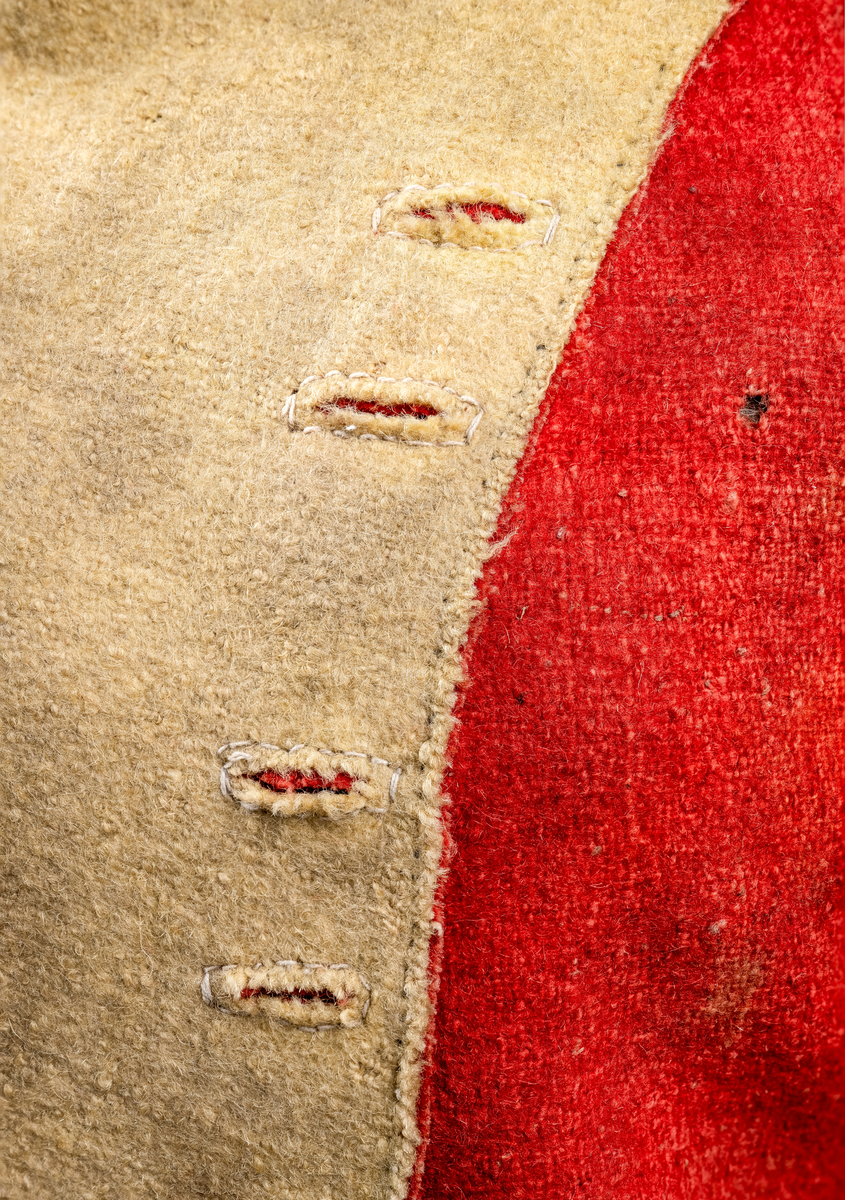 Kort trøye med høy ståkrage, sydd i grovt rødt lett valket ullstoff i empiresnitt. Trøya er helforet med grovt ullstoff. Kragen har mellomfor som er sydd fast til innerkragen. Ytterstoffet på kragen er brettet over på innsiden i øvre kant. Tittekant med rødt stoff i kortsidene på kragen. Trøya har søm MB og bakovertrekte skulder- og sidesømmer. Sidesømmene buer fra ermrigningen og mot MB. Sidesømmene slutter som legg med innlegg av gult stoff. Samme stoff brukes også som ytterkrage og slag på framstykket. Slagene er fastsydd og går opp som trekantforer mot kragen. Kneppes fast i bolen. 7 knapphull, men det mangler mange knapper. Plagget lukkes med 8 par metallhekter MF.  Nedenfor slagene er det påsydd trekanter av gråbeige ullstoff. Fra slagene foran skrår trøya bakover. Tosømsermer med dekor frampå ermene. Påsydd gult stoff med 2 striper av lys gråhvite bånd som går opp i en spiss mot innersømmen på ermet. Splitt ved yttersømmen. Denne lukkes med trekt rød knapp og knapphull. Mangler knappen på venstre erme. Stikninger langs slagene og kragen. Trøya er helt håndsydd og ytterkantene er sydd med sårkant på både for og stoff Tekst på 1 knapp, 3 knapper med stripe effekt inn mot midten.
Rød stoffhempe i nakken MB. Trøya har trolig tilhørt militæruniform fra tidlig 1800tall.