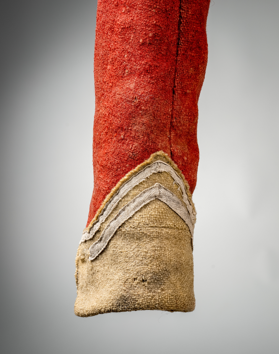 Kort trøye med høy ståkrage, sydd i grovt rødt lett valket ullstoff i empiresnitt. Trøya er helforet med grovt ullstoff. Kragen har mellomfor som er sydd fast til innerkragen. Ytterstoffet på kragen er brettet over på innsiden i øvre kant. Tittekant med rødt stoff i kortsidene på kragen. Trøya har søm MB og bakovertrekte skulder- og sidesømmer. Sidesømmene buer fra ermrigningen og mot MB. Sidesømmene slutter som legg med innlegg av gult stoff. Samme stoff brukes også som ytterkrage og slag på framstykket. Slagene er fastsydd og går opp som trekantforer mot kragen. Kneppes fast i bolen. 7 knapphull, men det mangler mange knapper. Plagget lukkes med 8 par metallhekter MF.  Nedenfor slagene er det påsydd trekanter av gråbeige ullstoff. Fra slagene foran skrår trøya bakover. Tosømsermer med dekor frampå ermene. Påsydd gult stoff med 2 striper av lys gråhvite bånd som går opp i en spiss mot innersømmen på ermet. Splitt ved yttersømmen. Denne lukkes med trekt rød knapp og knapphull. Mangler knappen på venstre erme. Stikninger langs slagene og kragen. Trøya er helt håndsydd og ytterkantene er sydd med sårkant på både for og stoff Tekst på 1 knapp, 3 knapper med stripe effekt inn mot midten.
Rød stoffhempe i nakken MB. Trøya har trolig tilhørt militæruniform fra tidlig 1800tall.