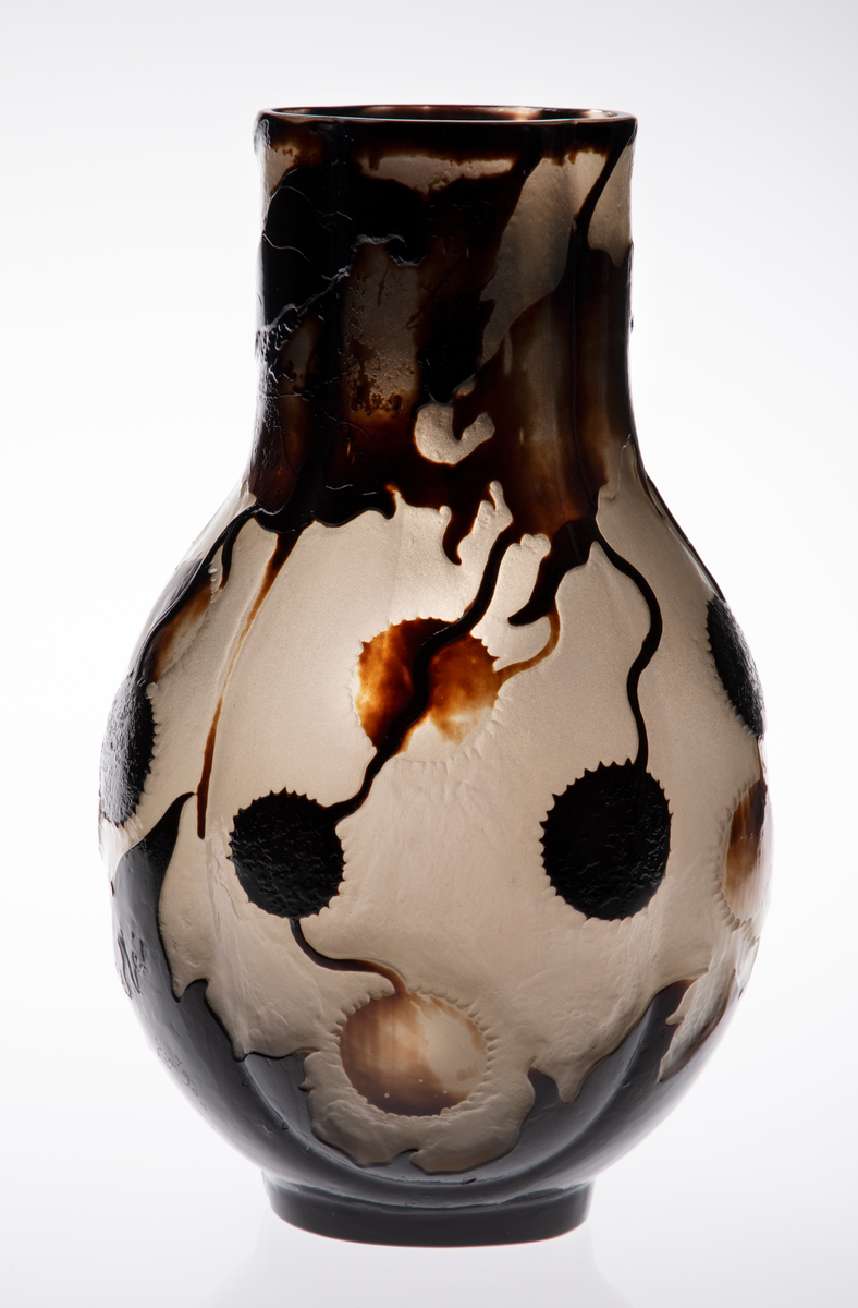 Glassvase med vid hals. Oval utforming med konvekse sider på en sirkulær fot. Overflaten består av en nesten opak gråhvit glassmasse, som er dekorert med blomster og blader fra platantreet i silhuett. Dekorfargen er gradert fra lysebrun til nærmest sort. Signert ”Gallé – Ex. 1900”.