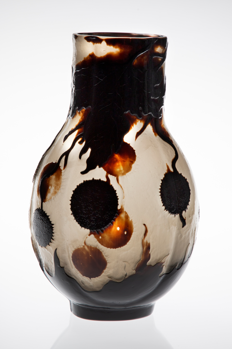 Glassvase med vid hals. Oval utforming med konvekse sider på en sirkulær fot. Overflaten består av en nesten opak gråhvit glassmasse, som er dekorert med blomster og blader fra platantreet i silhuett. Dekorfargen er gradert fra lysebrun til nærmest sort. Signert ”Gallé – Ex. 1900”.