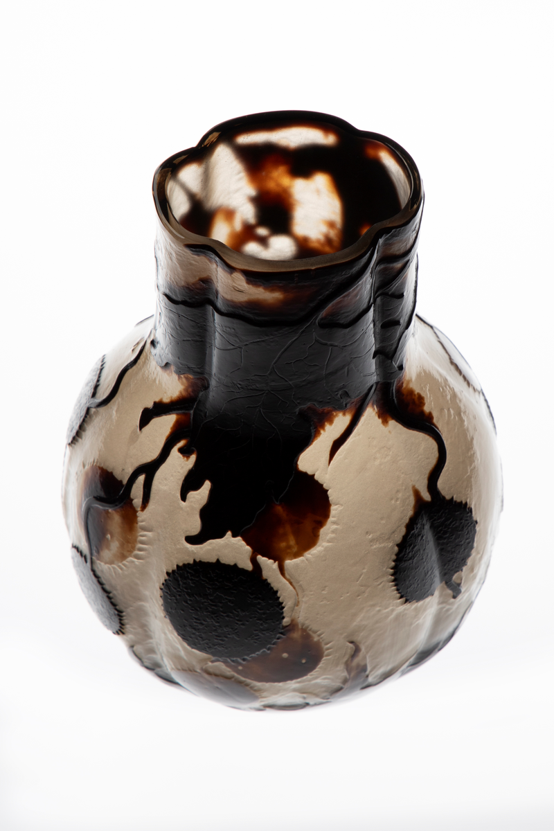 Glassvase med vid hals. Oval utforming med konvekse sider på en sirkulær fot. Overflaten består av en nesten opak gråhvit glassmasse, som er dekorert med blomster og blader fra platantreet i silhuett. Dekorfargen er gradert fra lysebrun til nærmest sort. Signert ”Gallé – Ex. 1900”.