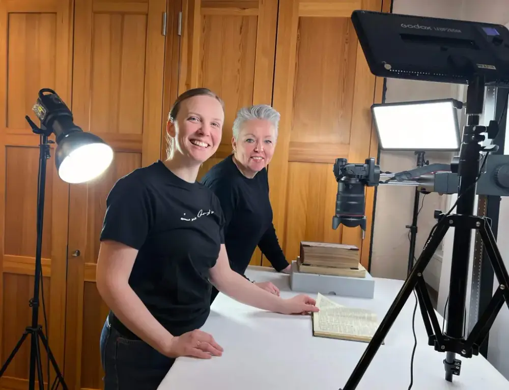 Stine Stennes Hovdenakk og Siri Beate Gjerde smiler mot fotografen ved høg arbeidsbenk med bøker, kamera og lampar. 