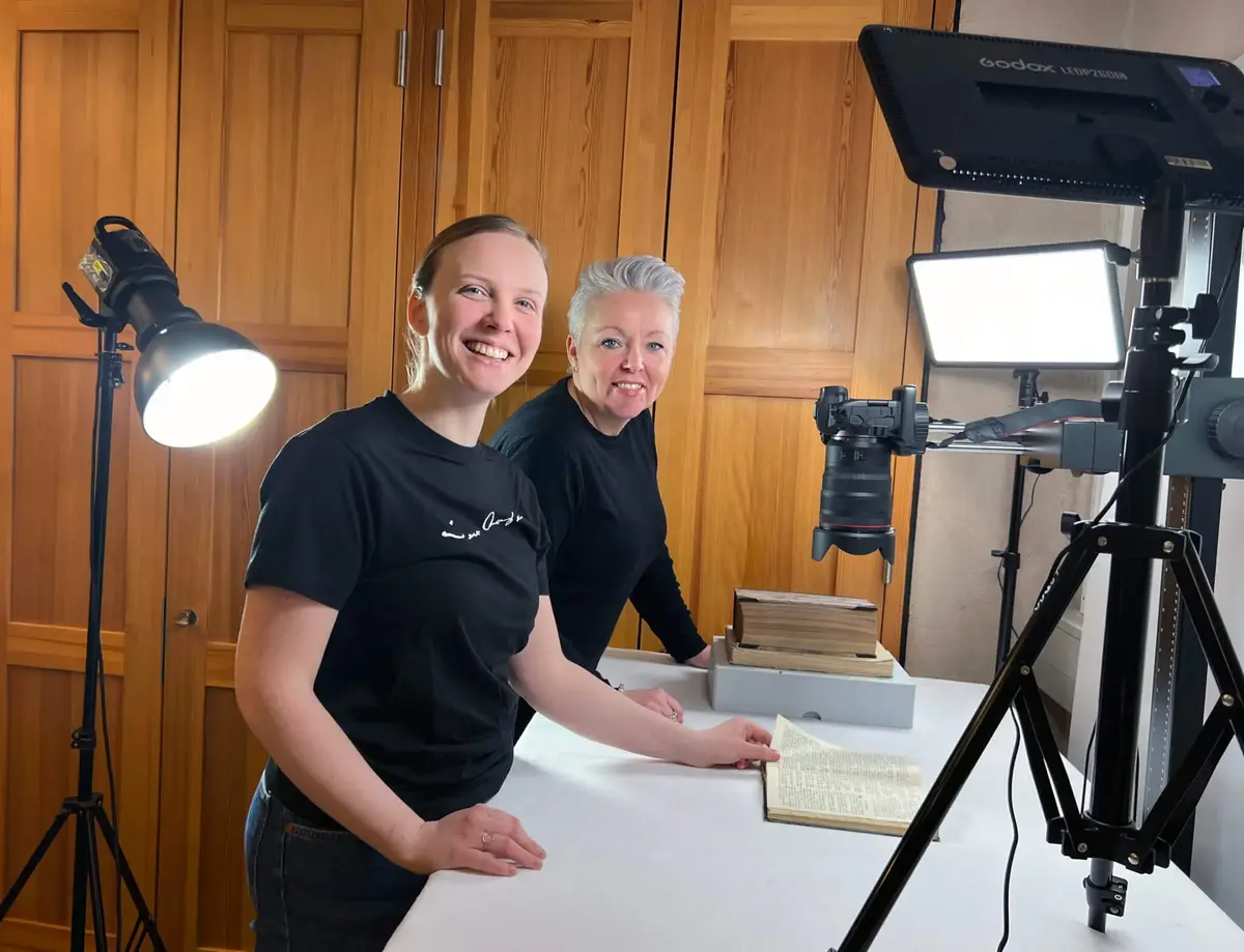 Stine Stennes Hovdenakk og Siri Beate Gjerde smiler mot fotografen ved høg arbeidsbenk med bøker, kamera og lampar. 