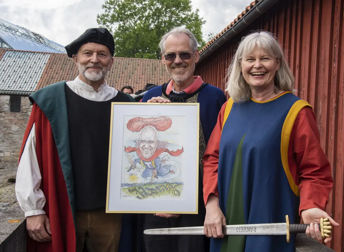 To menn og en dame i fargerike middelalderklær. Mann i blå kappe holder en karrikaturtegning av seg selv. Han er Mesterborger av Hammer By. Kvinnen holder et sverd og er ordfører i Hamar.