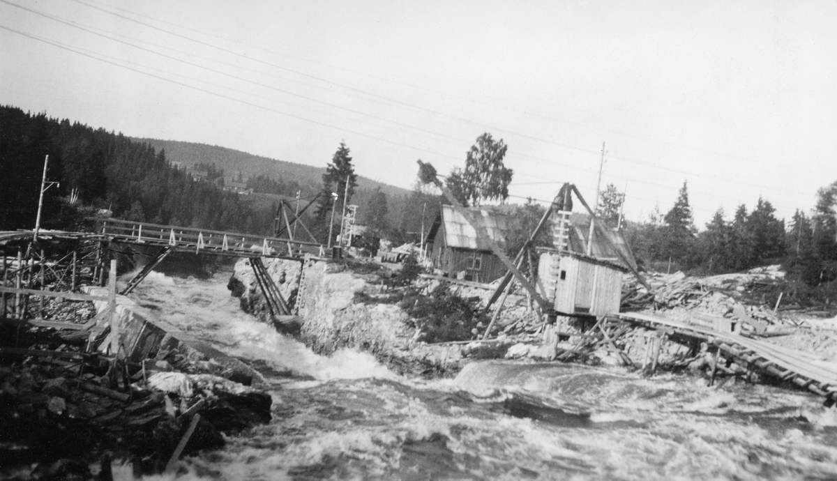 Fra tømmerkanalen ved Vamma i Askim i Østfold (nedre Glomma).  Denne kanalen ble innviet i 1927, etter at Fredrikstad Tømmerdirektion i elleve sesonger (1916-26) med vekslende hell hadde forsøkt å fløte de store tømmermengdene i denne delen av vassdraget gjennom ei underdimensjonert tømmerrenne litt lengre vest.  Fotografiet viser hvordan en del av kanalen var nedsprengt i berggrunnen i tolv meters bredde.  Da dette fotografiet ble tatt sto en del av installasjonene som ble brukt under kanalbygginga fortsatt intakte.  Til høyre ser vi ei slags kjørebane med skinnelegging, antakelig for vogner ("vagger") som ble brukt i forbindelse med transport av materiell.  Ved denne transportbanen ser vi også et lite bordskur med pulttak, og i forkant av dette er det ei kran ut mot kanalløpet.  Lengre nede er det bygd ei bru over kanalen, mot en bygning på høyre bredd.  Da dette fotografiet ble tatt var strømmen i kanalen - etter skumtoppene å dømme - meget stri. 