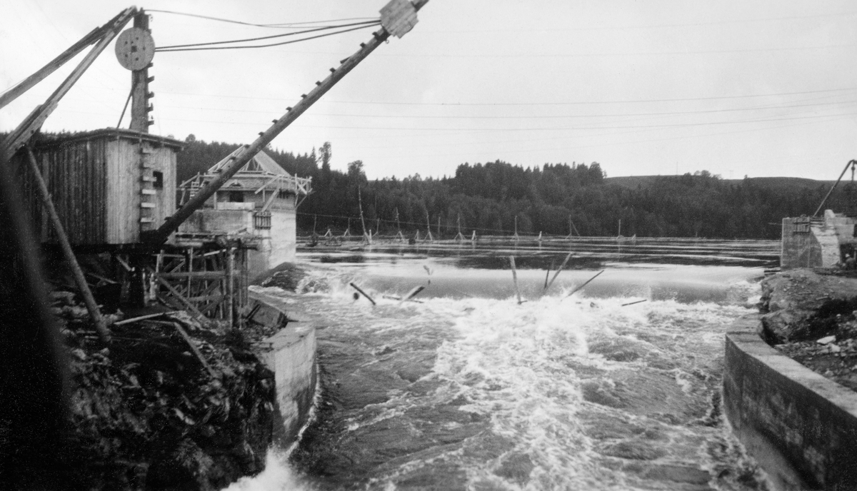 Fra tømmerkanalen ved Vamma i Askim i Østfold (nedre Glomma).  Denne kanalen ble innviet i 1927, etter at Fredrikstad Tømmerdirektion i elleve sesonger (1916-26) med vekslende hell hadde forsøkt å fløte de store tømmermengdene i denne delen av vassdraget gjennom ei underdimensjonert tømmerrenne litt lengre vest.  Fotografiet er antakelig tatt fra ei bru over kanalen (jfr. SJF. 1989-04439) oppover mot det 35 meter brede innløpet til den nye kanalen.  Vi der at vannet fra kraftverksdammen passerer en terskel - som egentlig var ei sektorluke som kunne heves og senkes over ei gruve i kanalbotnen.  Nedenfor innløpet smalner kanalen til tolv meters bredde.  I dette partiet valgte ingeniørene som sto bak dette prosjektet å skjerme kanalbreddene med loddrette betongmurer.  På den venstre bredden ser vi et treskur med ei slags kran, som antakelig ble brukt i arbeidet med kanalen.  Lengre bak skimter vi et hus som ble bygd på karet ved den ene sida av kanalinnløpet.  Det er nærliggende å anta at det var herfra at posisjonen til den nevnte sektorluka ble regulert for å sikre riktig vannføring i kanalen, uansett vannstand i kraftverksdammen. 