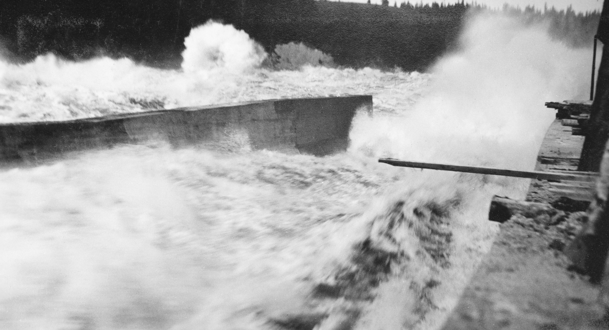 Fra tømmerkanalen ved Vamma i Askim i Østfold (nedre Glomma).  Denne kanalen ble innviet i 1927, etter at Fredrikstad Tømmerdirektion i elleve sesonger (1916-26) med vekslende hell hadde forsøkt å fløte de store tømmermengdene i denne delen av vassdraget gjennom ei underdimensjonert tømmerrenne litt lengre vest.  Fotografiet er tatt nederst i den cirka 360 meter lange kanalen, som i dette partiet var forholdsvis bratt (1:3).  På grunn av hellingsgraden hadde ingeniørene som planla den nye kanalen funnet det nødvendig å gi den nedre delen forholdsvis høye, loddrette sidevegger av betong, og disse er godt synlige på dette fotografiet.  Vi ser også at kanalen fikk noe av en ilddåp, i og med at den første driftssesongen, 1927, falt sammen med et av de betydeligste flomårene i Glomma på 1900-tallet.  Fotografiet viser at både tømmerkanalen og hovedløpet gikk flomstore og kvitskummende. 