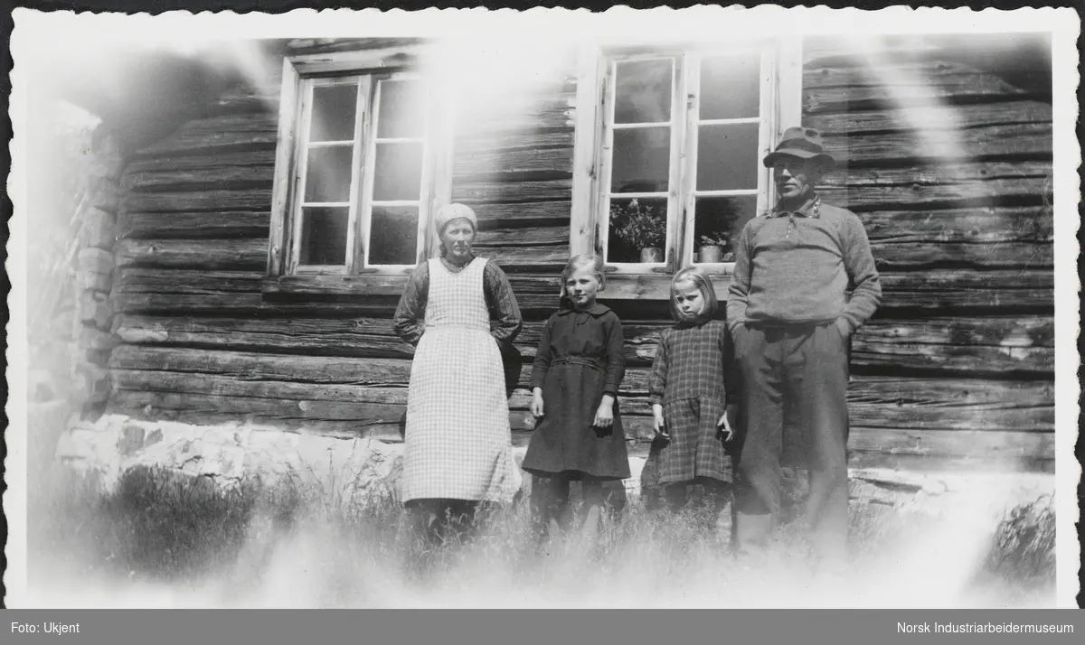 Mann og kvinne med to barn mellom seg stående foran laftet bolig.