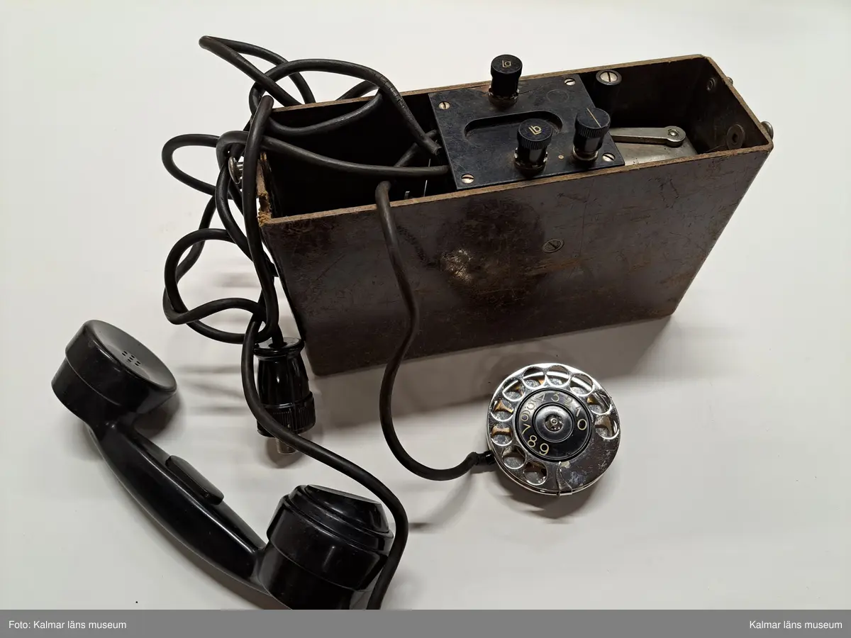 KLM 46809. Telefon, portativtelefon, fälttelefon. Fyrkantig låda av brunt trä och lock med metallspännen, Inuti lådan lur av svart bakelit, nummerskiva av metall och svart gummisladd. I mittsektionen tre knappar, varav två med bokstäverna: Lb resp. La. På nummerskivans kant tillverkarstämpel: Telefon A.-B. LM Ericsson Stockholm 12 51. PÅ lockets ovansida text: Ericsson.