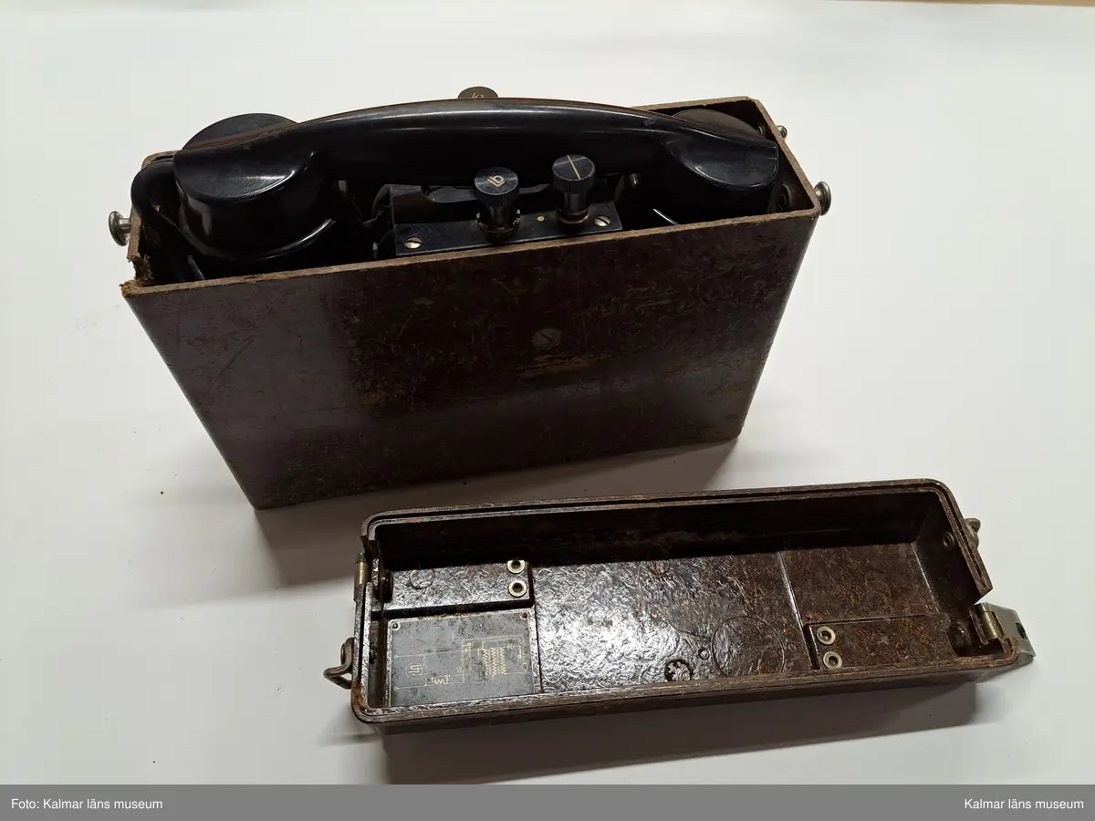 KLM 46809. Telefon, portativtelefon, fälttelefon. Fyrkantig låda av brunt trä och lock med metallspännen, Inuti lådan lur av svart bakelit, nummerskiva av metall och svart gummisladd. I mittsektionen tre knappar, varav två med bokstäverna: Lb resp. La. På nummerskivans kant tillverkarstämpel: Telefon A.-B. LM Ericsson Stockholm 12 51. PÅ lockets ovansida text: Ericsson.