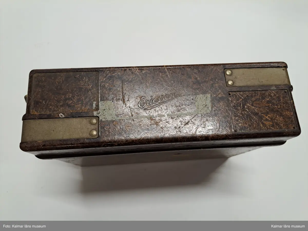 KLM 46809. Telefon, portativtelefon, fälttelefon. Fyrkantig låda av brunt trä och lock med metallspännen, Inuti lådan lur av svart bakelit, nummerskiva av metall och svart gummisladd. I mittsektionen tre knappar, varav två med bokstäverna: Lb resp. La. På nummerskivans kant tillverkarstämpel: Telefon A.-B. LM Ericsson Stockholm 12 51. PÅ lockets ovansida text: Ericsson.