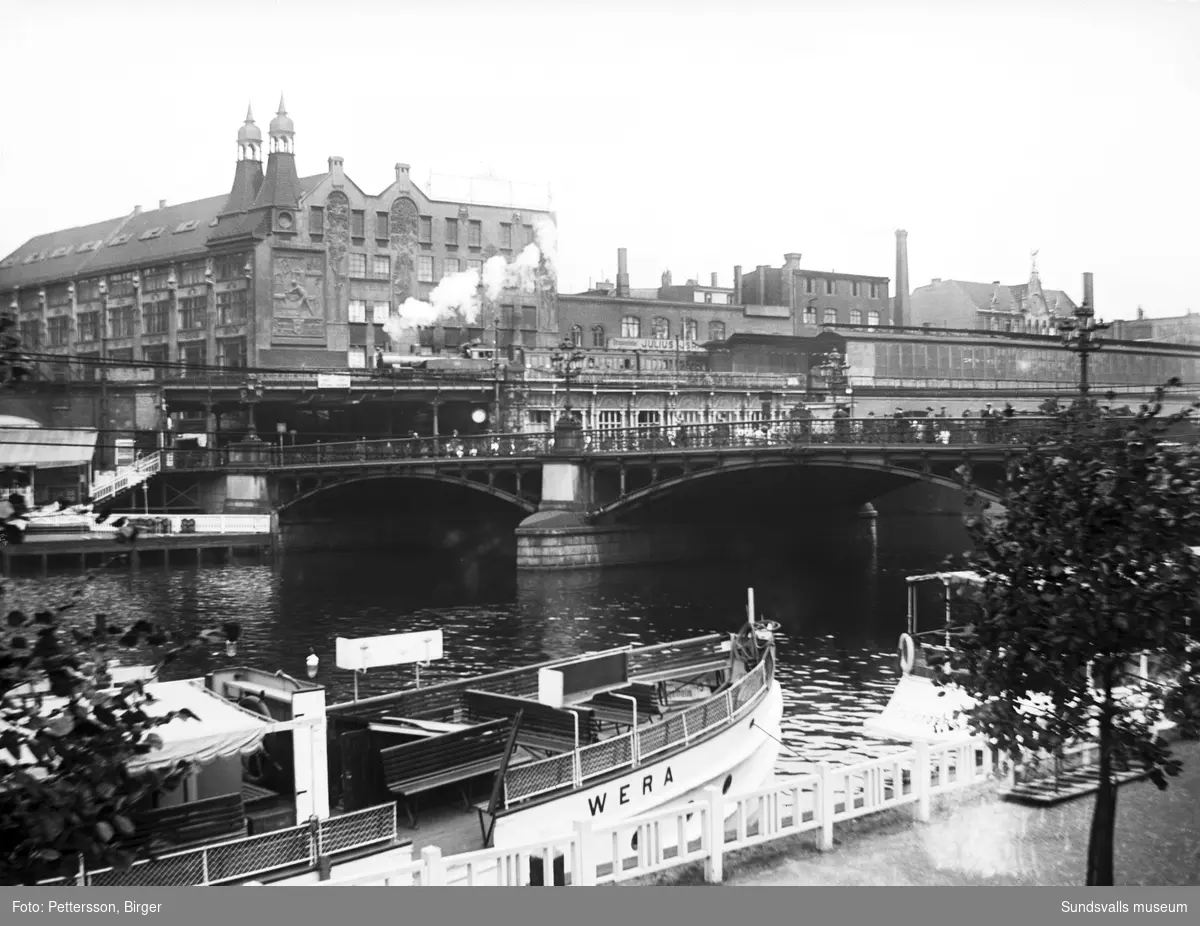 Stadsvy i Berlin, en bro över Spree och vid kajen en passagerarbåt vid namn Wera. På andra sidan en fabriksbyggnad med vacker fasaddekor.