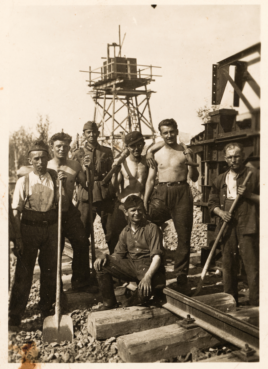 Gruppebilde av tsjekkiske tvangsarbeidere og en tysk soldat, tatt på en anleggsplass. Et slags tårn i bakgrunnen.