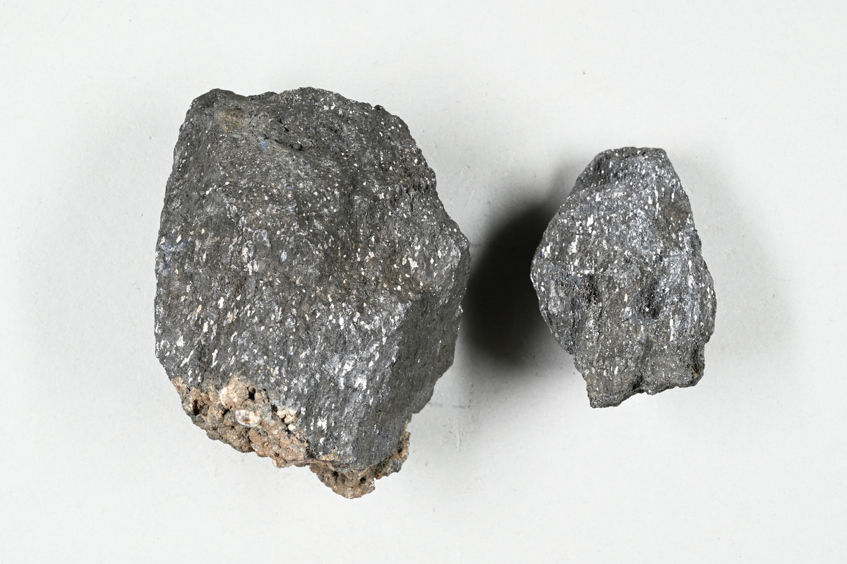 Två materialprov, grå och brunaktig. Metalliskt skimmer i båda. Tillhörande lapp med notering: "Ugnsbrott (?), från Siljanhyttan (?). Falun".