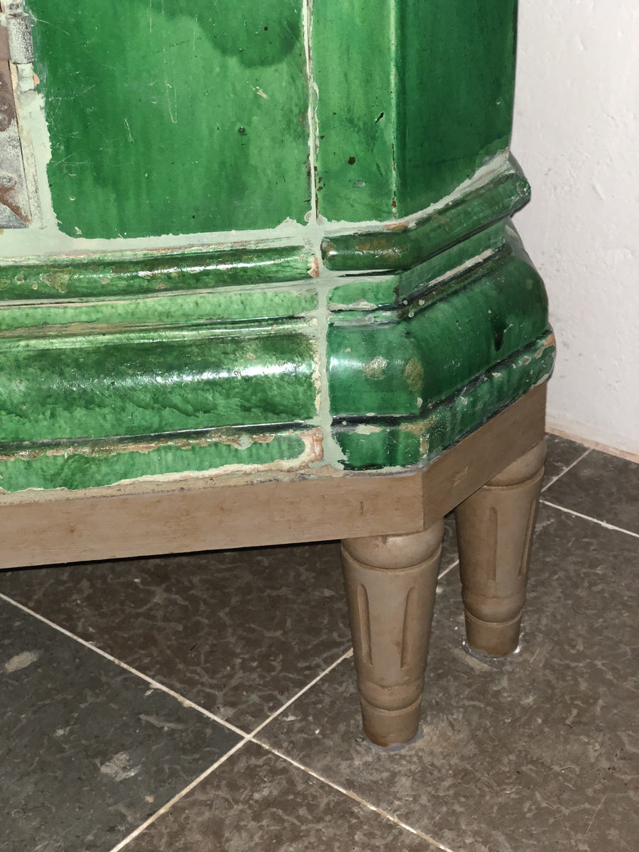 Kakelugn flat, av lergods med grön, lite flammig blyglasyr. Fasade hörn, rakt avslutande upptill med vulstformat kakel, nedtill konvext simskakel. Uppsatt i slät modell utan skänkhylla på fotställning i gustaviansk stil.