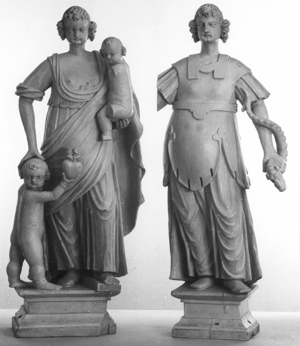 Kyrklig skulptur, trä, staty från gamla korskranket i Heliga Trefaldighets kyrka i Gävle, av Ewert Friis omkring 1665. 

Stående kvinnofigur med naket barn på vänster arm samt stående naket barn vid höger sida. Sistnämnda barn har ett brinnande hjärta i höger hand och kvinnans vänstra hand på sitt huvud. Vänster ben är främst, foten avslagen. Sittande barnets ena ben och andra fot avslagna. 
Fyrsidigt, profilerat postament. 
Gråvit färg.
