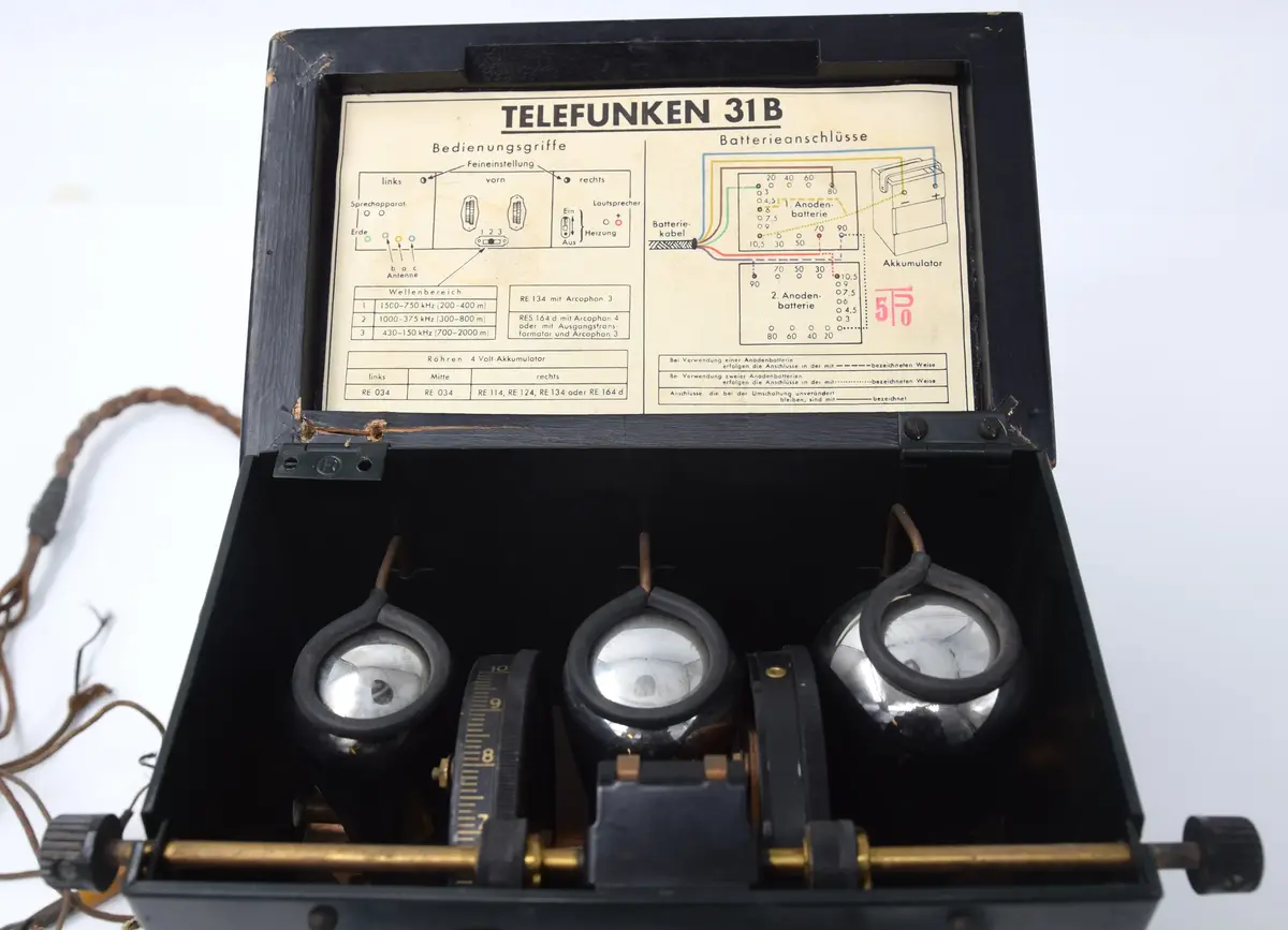 Radioapparat, för batterianslutning, av märke: Telefunken. Låda av mörkgrön plåt med lock av svartmålat trä, och fotplatta av trä. På baksidan firmamärke: Telefunken 31 B No. 43516. På locket skylt med: Telefunken. I vänster sida: 6 hål för antenner och i höger sida 2 hål samt knapp märkt: EIN. På framsidan en spak, rörlig i sidled från: 1,2,3. samt två trissor, numrerade 0-10, manövrerade från 2 skruvar i sidornas överkant. På baksidan fästad en sladd, delad i 6 delar. I locket fäst en pappersetikett med bruksanvisning.