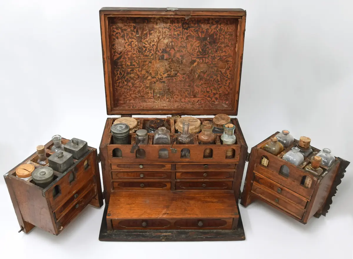 Medikamentlåda, av lövträ. Rektangulärt skrin, som är limmat samman. Försett med kupigt lock på gångjärn. På locket ett handtag av en järnten. Gångjärnen är förlängda till beslag i silverfärg liknande växtornament. Liknande beslag finns på framsidan och runt nyckelhålen. I hörnen finns beslag liknande blommor. Beslagen är gjorda av 1,6 mm tjock plåt. Stängs med nyckel samt hakar på lockets sidor. Främre delen av skrinet är uppdelad i två delar som kan svängas ut åt sidorna på gångjärn på skrinets sidor. Skrinet är uppdelat i tre våningar: den översta består av kubiska fack, var i glasflaskor och glas och plåtburkar med medicin i. Andra våningen består av små lådor klädda inuti med papper. Understa våningen består av en stor låda innehållande en våg av järn med skålar av guttaperka. Insidan av locket är klätt med papper med växtrankor och i mitten inom en ram två krönta örnar under en större krona.