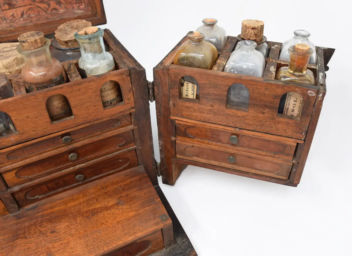 Medikamentlåda, av lövträ. Rektangulärt skrin, som är limmat samman. Försett med kupigt lock på gångjärn. På locket ett handtag av en järnten. Gångjärnen är förlängda till beslag i silverfärg liknande växtornament. Liknande beslag finns på framsidan och runt nyckelhålen. I hörnen finns beslag liknande blommor. Beslagen är gjorda av 1,6 mm tjock plåt. Stängs med nyckel samt hakar på lockets sidor. Främre delen av skrinet är uppdelad i två delar som kan svängas ut åt sidorna på gångjärn på skrinets sidor. Skrinet är uppdelat i tre våningar: den översta består av kubiska fack, var i glasflaskor och glas och plåtburkar med medicin i. Andra våningen består av små lådor klädda inuti med papper. Understa våningen består av en stor låda innehållande en våg av järn med skålar av guttaperka. Insidan av locket är klätt med papper med växtrankor och i mitten inom en ram två krönta örnar under en större krona.