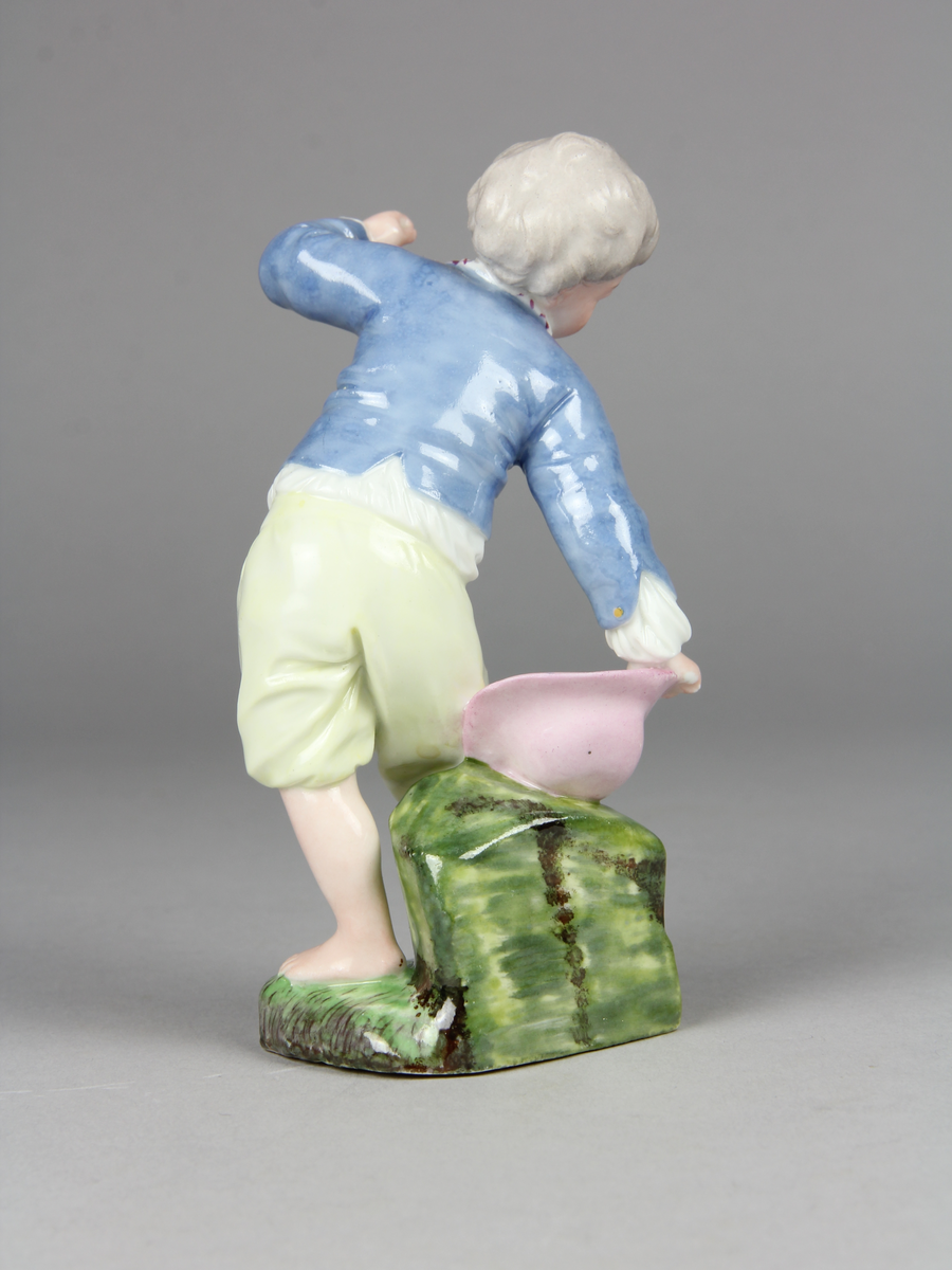 Pojke bär en hatt med ägg i och tappar den. Han verkar ha tappat hattens ena brätte, viker sig åt sin högra sida, håller upp vänster arm och har ett häpet ansiktsuttryck. På marken under hatten ligger ett sönderslaget ägg.