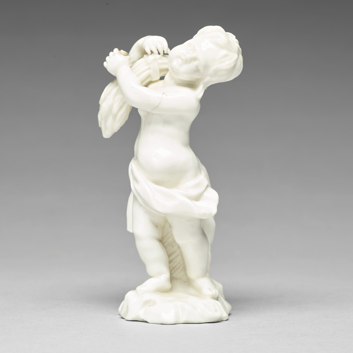 Figurin föreställande putto (italienska för "liten pojke") som bär en sädeskärve. Kring höften hänger en draperad textil. Bakstycke och fotplatta med draperad textil.