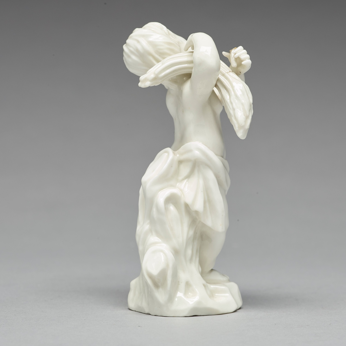 Figurin föreställande putto (italienska för "liten pojke") som bär en sädeskärve. Kring höften hänger en draperad textil. Bakstycke och fotplatta med draperad textil.