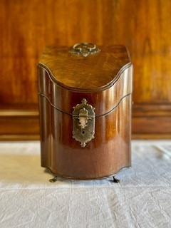 Flaskschatull fanerat med mahogny, vilande på fötter av mässing. Schatullet har ett slutande lock som möter upp i en en konvex framsida. På locket ett handtag av mässing. Raka sidor med handtag av mässing. Mitt fram en låsanordning med en profilerad bricka. Schatullets insida är uppdelad i tre fack, alla fodrad med sammet i en röd och grön nyans.  Fodrets sömn är täckt med kantband i guld. Insidans fack är anpassade för tre flaskor var av två i fyrkantig modell och en i bågformad modell. 