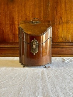 Flaskschatull fanerat med mahogny, vilande på fötter av mässing. Schatullet har ett slutande lock som möter upp i en en konvex framsida. På locket ett handtag av mässing. Raka sidor med handtag av mässing. Mitt fram en låsanordning med en profilerad bricka. Schatullets insida är uppdelad i tre fack, alla fodrad med sammet i en röd och grön nyans.  Fodrets sömn är täckt med kantband i guld. Insidans fack är anpassade för tre flaskor var av två i fyrkantig modell och en i bågformad modell. 