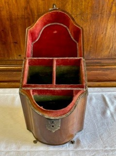 Flaskschatull fanerat med mahogny, vilande på fötter av mässing. Schatullet har ett slutande lock som möter upp i en en konvex framsida. På locket ett handtag av mässing. Raka sidor med handtag av mässing. Mitt fram en låsanordning med en profilerad bricka. Schatullets insida är uppdelad i tre fack, alla fodrad med sammet i en röd och grön nyans.  Fodrets sömn är täckt med kantband i guld. Insidans fack är anpassade för tre flaskor var av två i fyrkantig modell och en i bågformad modell. 