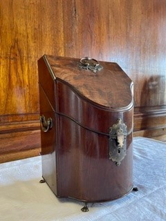 Flaskschatull fanerat med mahogny, vilande på fötter av mässing. Schatullet har ett slutande lock som möter upp i en en konvex framsida. På locket ett handtag av mässing. Raka sidor med handtag av mässing. Mitt fram en låsanordning med en profilerad bricka. Schatullets insida är uppdelad i tre fack, alla fodrad med sammet i en röd och grön nyans.  Fodrets sömn är täckt med kantband i guld. Insidans fack är anpassade för tre flaskor var av två i fyrkantig modell och en i bågformad modell. 