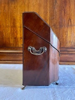Flaskschatull fanerat med mahogny, vilande på fötter av mässing. Schatullet har ett slutande lock som möter upp i en en konvex framsida. På locket ett handtag av mässing. Raka sidor med handtag av mässing. Mitt fram en låsanordning med en profilerad bricka. Schatullets insida är uppdelad i tre fack, alla fodrad med sammet i en röd och grön nyans.  Fodrets sömn är täckt med kantband i guld. Insidans fack är anpassade för tre flaskor var av två i fyrkantig modell och en i bågformad modell. 
