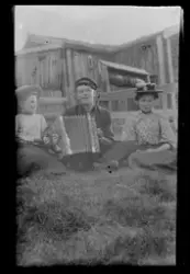 Tre personer sittende foran hus. En med trekkspill.