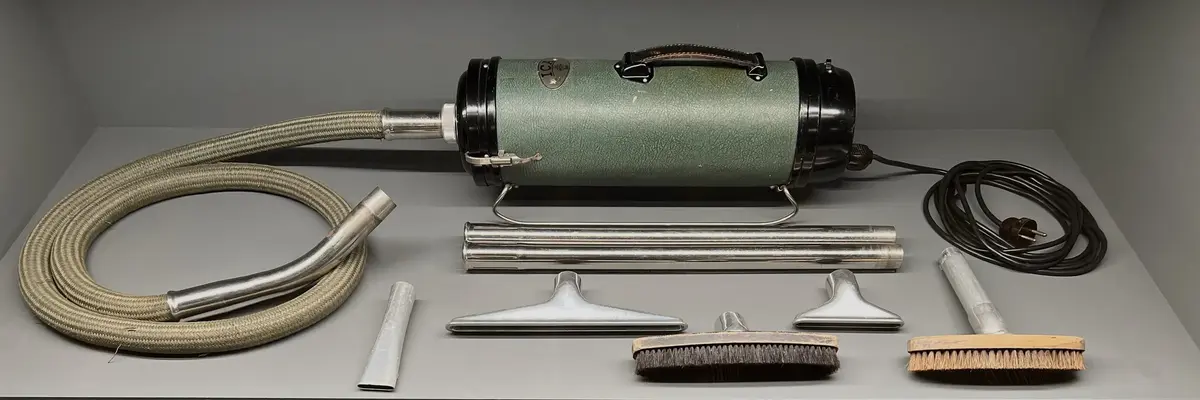 Grön dammsugare med diverse munstycken och tillbehör. Totalt 10 delar. Dammsugaren har en grön cylindriskt kropp med två metallben som sträcker sig över dammsugarens längd. Läderhandtag på dammsugarens ovansida. 
Dammsugarens slag klädd med grön flätad textil. Svart hängare i metall för upphängning  på vägg av dammsugaren. 