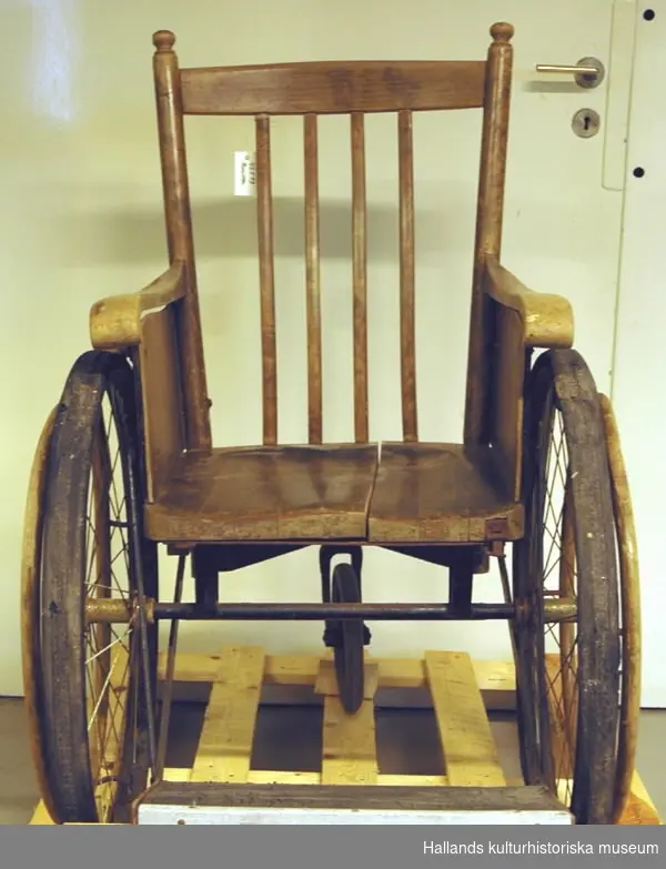 Rullstol i trä med tre hjul, två stora och ett mindre. Märkt på baksidan "KSA6an".