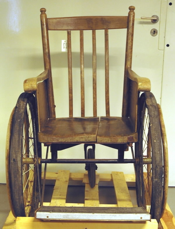 Rullstol i trä med tre hjul, två stora och ett mindre. Märkt på baksidan "KSA6an".