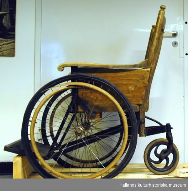 Rullstol i trä med tre hjul, två stora och ett mindre. Märkt på baksidan "KSA6an".